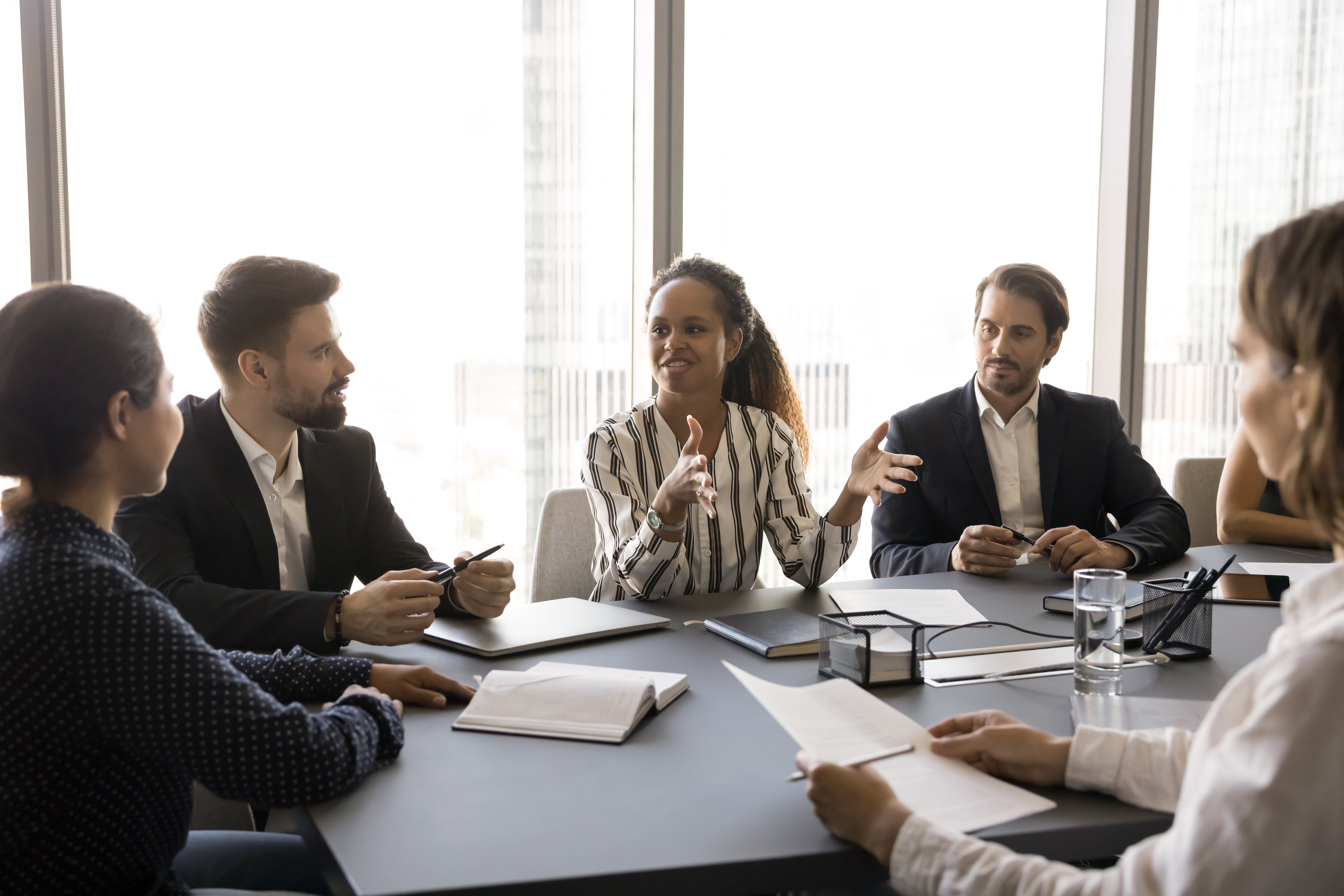 Five people sit around a conference table in a modern office, engaged in a discussion about AI in private equity. One woman in the center gestures as she speaks, while others listen attentively. Papers, notebooks, and glasses of water are on the table.