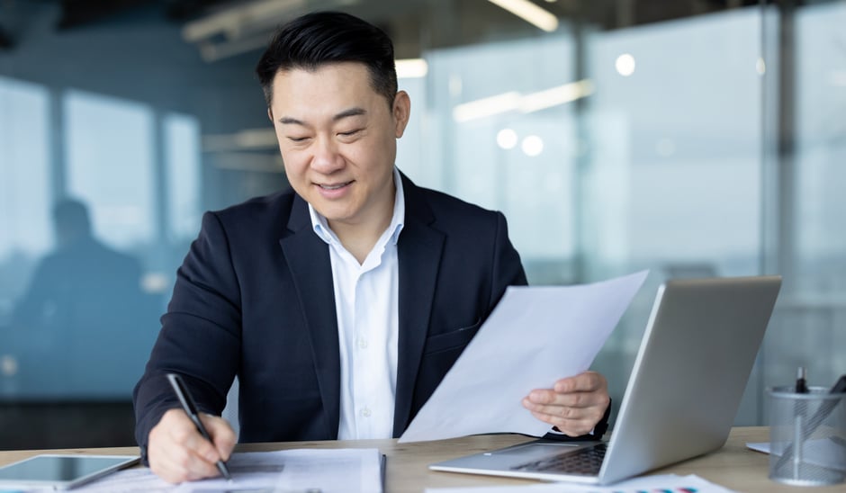A man in a suit sits at a desk, smiling while holding papers and writing. With documents and a laptop nearby, he appears to be working in a modern office, possibly analyzing the impact of ai in private equity through large windows.