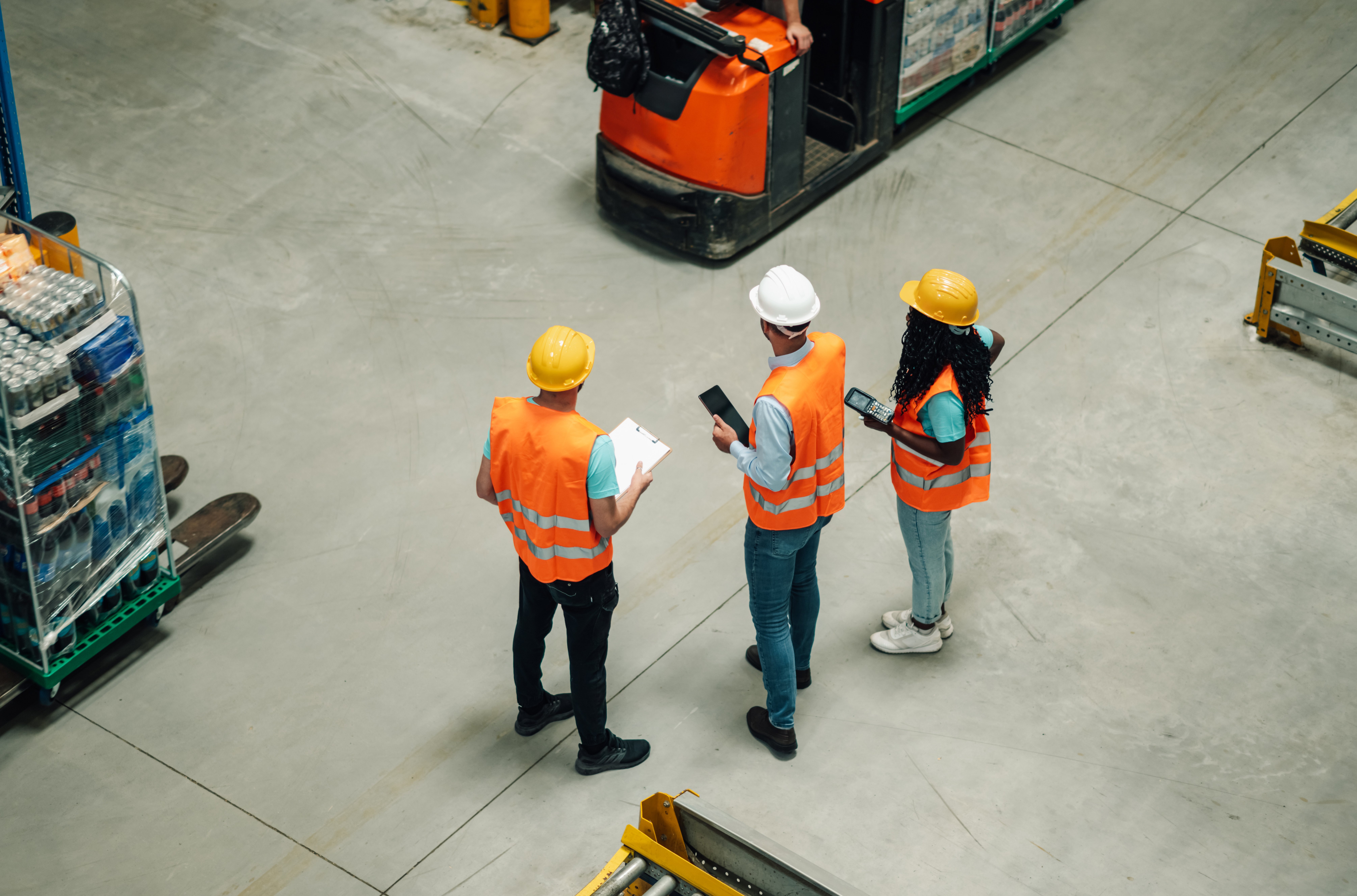 Three workers in safety vests and helmets stand in a warehouse, discussing supply chain AI while holding a clipboard and tablet. Pallets with goods are nearby, and a forklift is visible in the background.