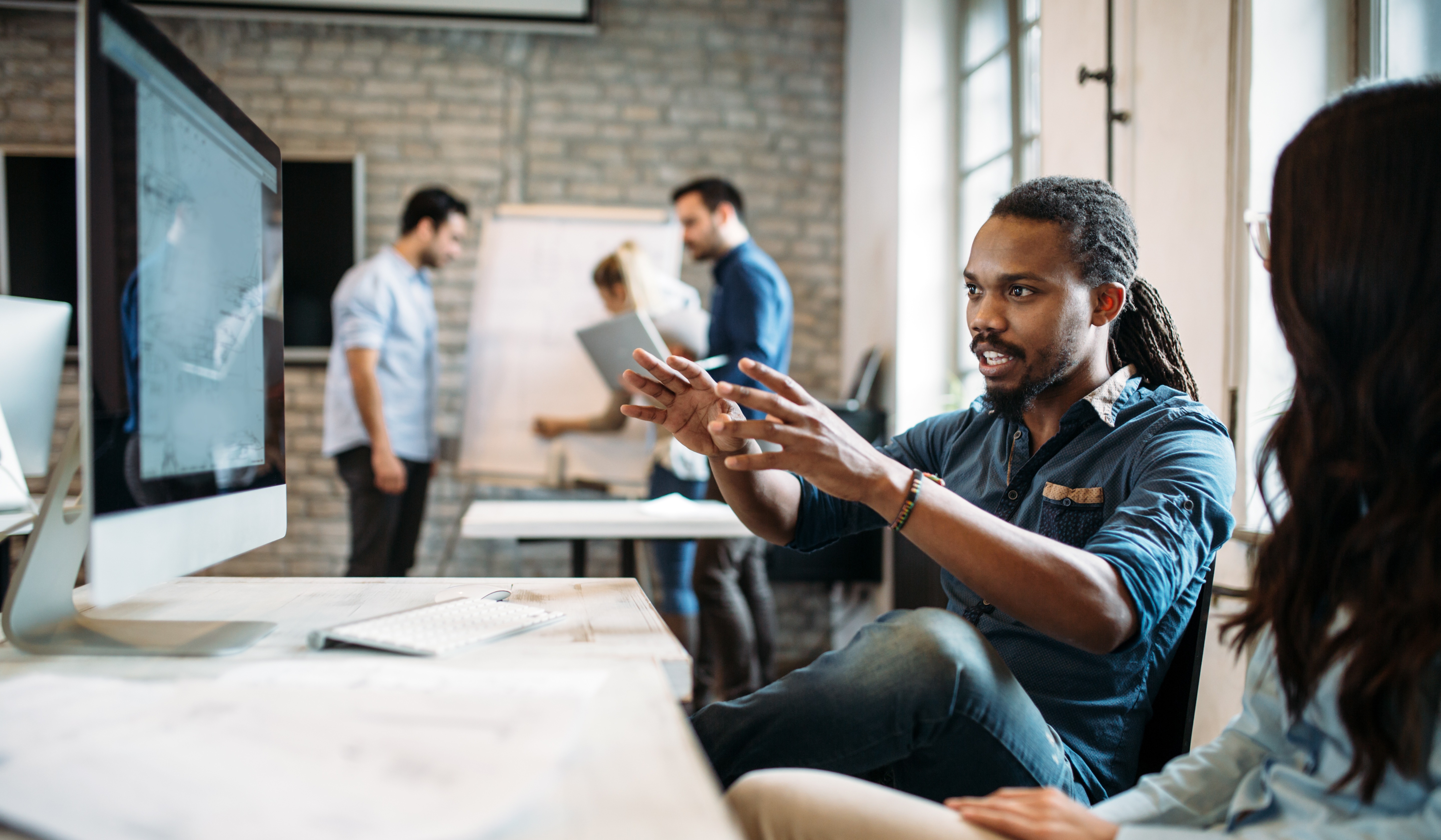 Business team collaborating on ERP modernization strategy. A man explains ideas at a computer while colleagues discuss at a whiteboard in the background, representing how organizations plan composable ERP solutions to replace outdated systems.