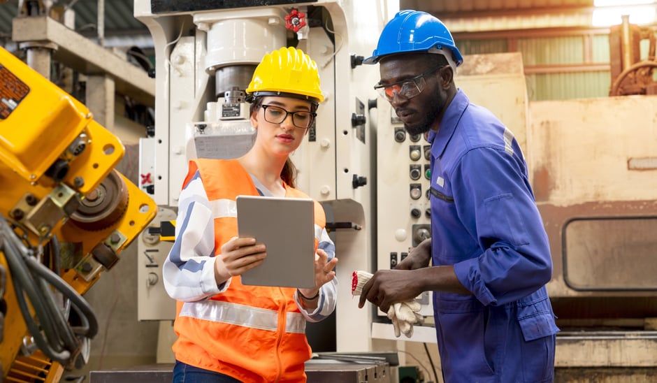 A woman in a yellow hard hat and orange safety vest holds a tablet, discussing data integration challenges with a man in a blue hard hat and work attire. They stand in an industrial setting, surrounded by machinery.