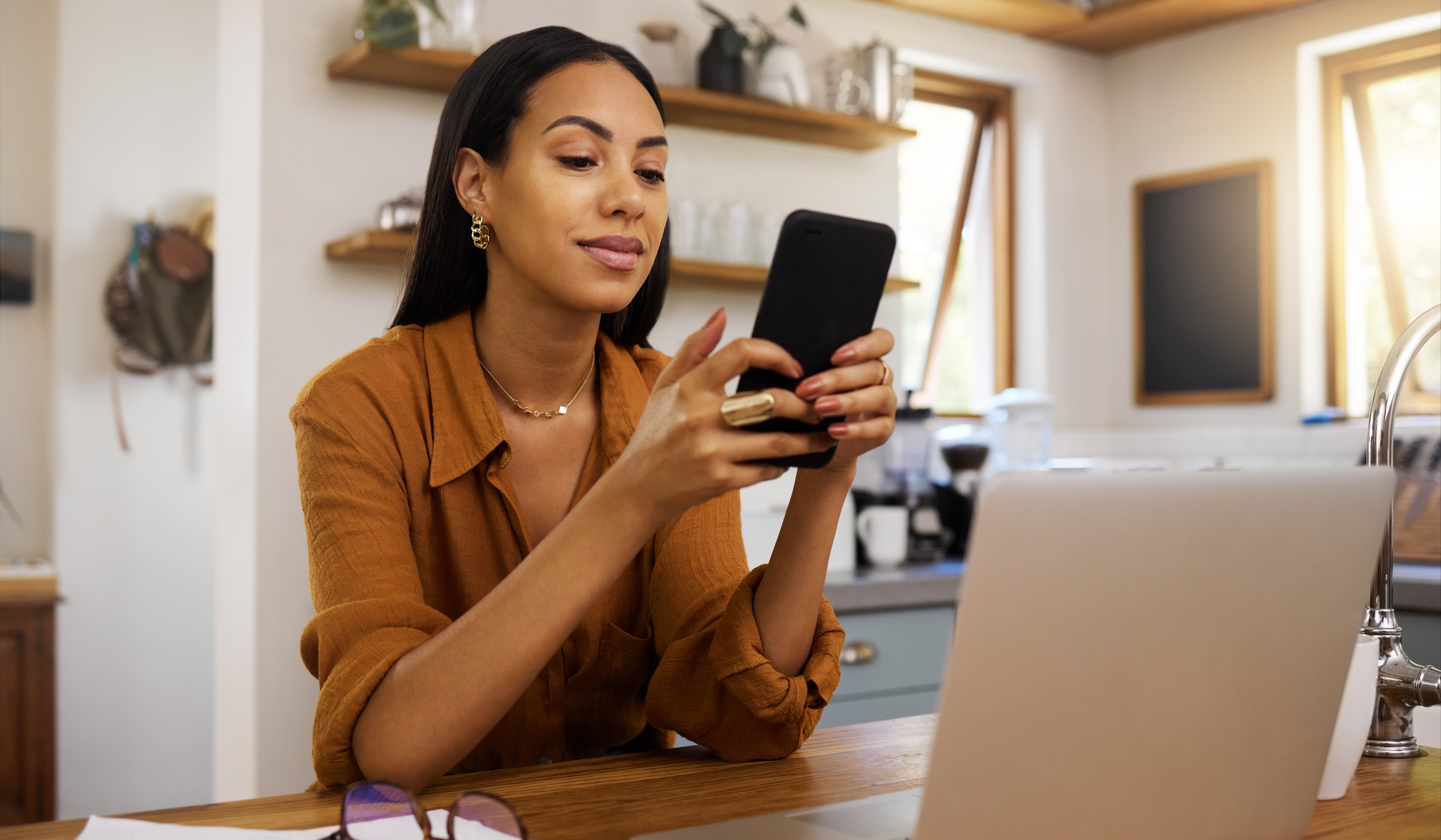 A woman in a brown shirt sits at a kitchen table, smiling at her smartphone, likely reviewing app development ROI. She has a laptop open in front of her and eyeglasses beside it. Shelves with jars and a window create a cozy background.