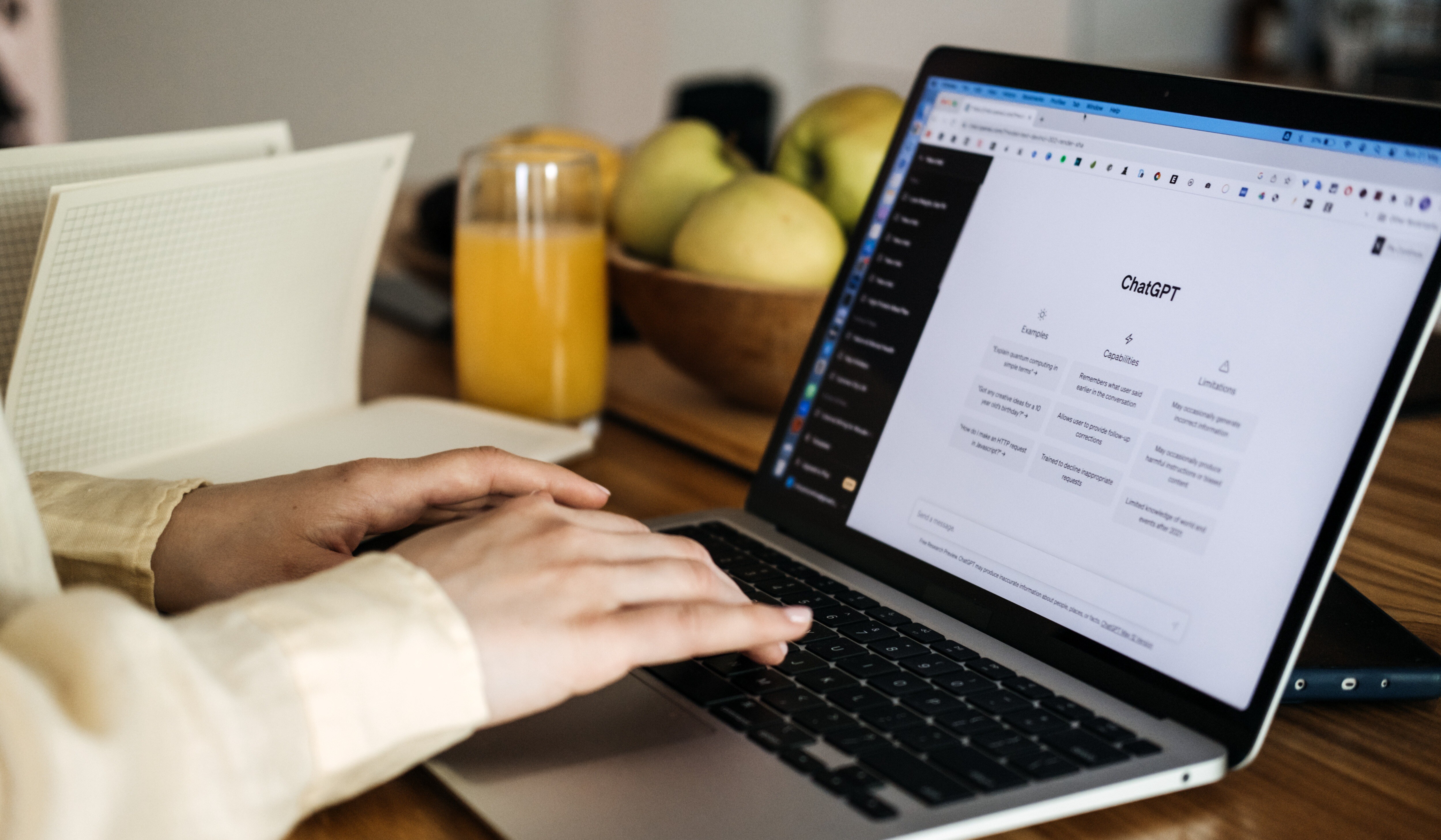 A person is typing on a laptop displaying the ChatGPT webpage, exploring enterprise AI applications. A bowl of apples and a glass of orange juice rest on the wooden table beside the laptop, while a notebook is partially visible in the background.