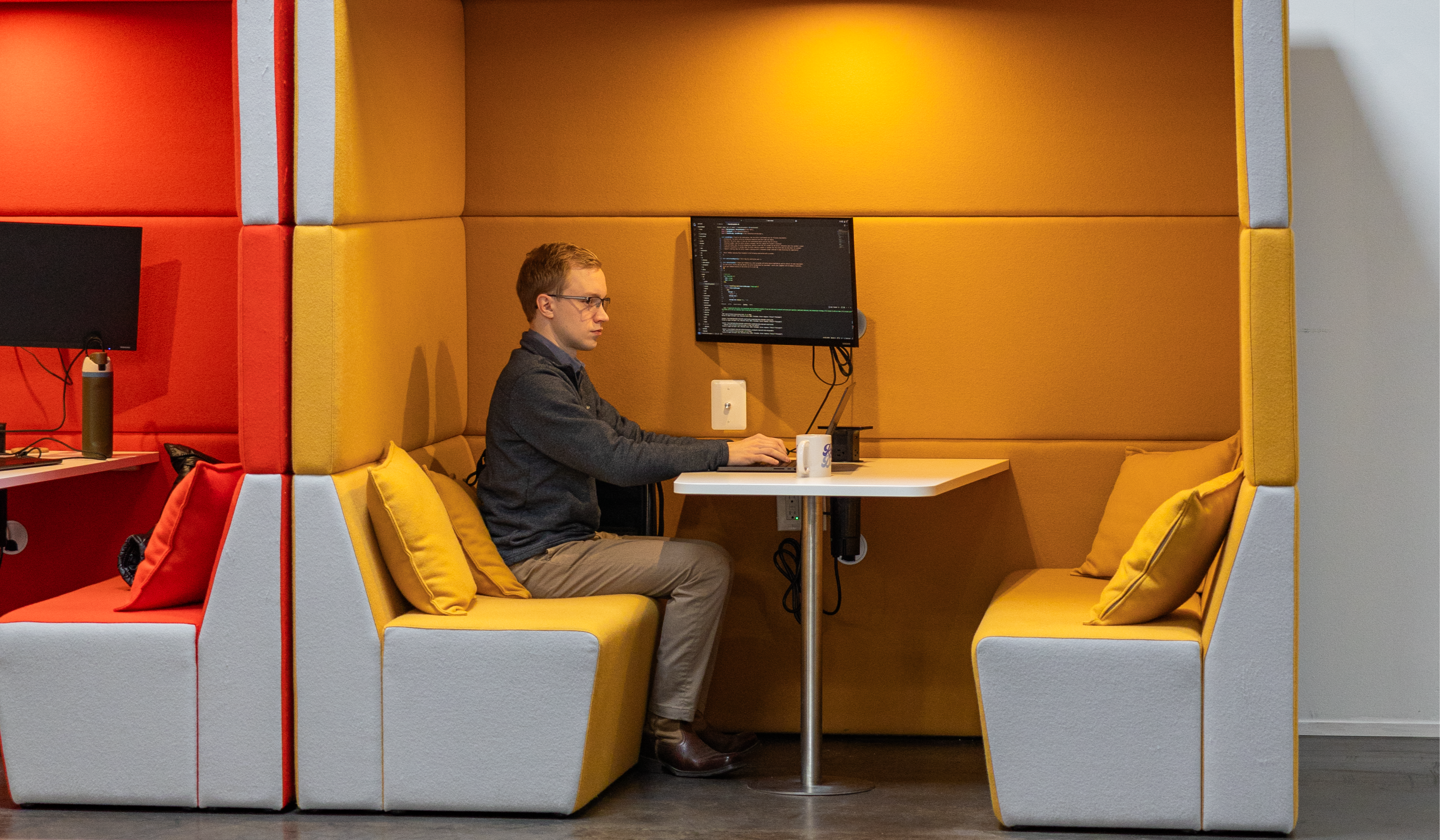 A man sits at a small table inside a yellow and orange padded booth, working on enterprise AI code displayed on his computer screen. He is focused, with a coffee mug beside him on the desk.