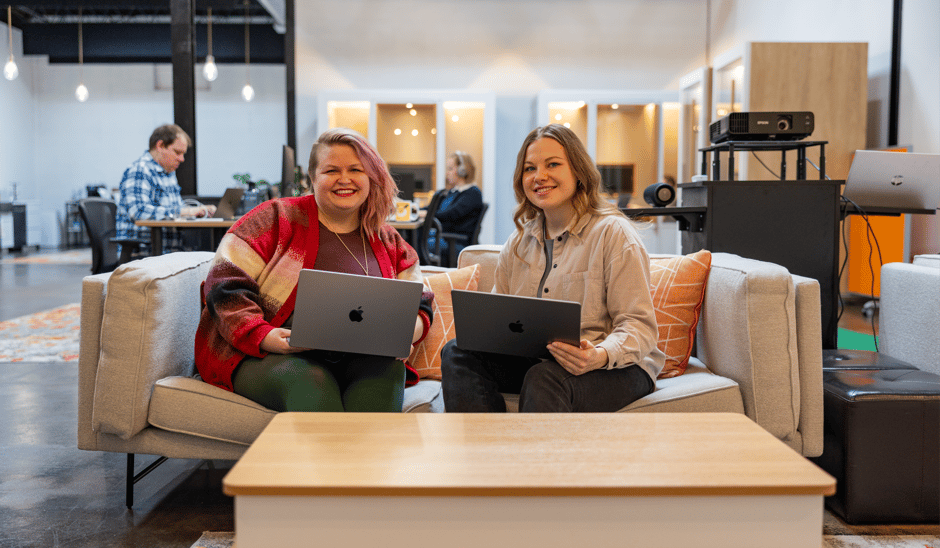 Two people sitting on a gray sofa with laptops, smiling at the camera in a modern office environment focused on cross-platform development. A man is working at a desk in the background, and the room has an open, airy feel with warm lighting.