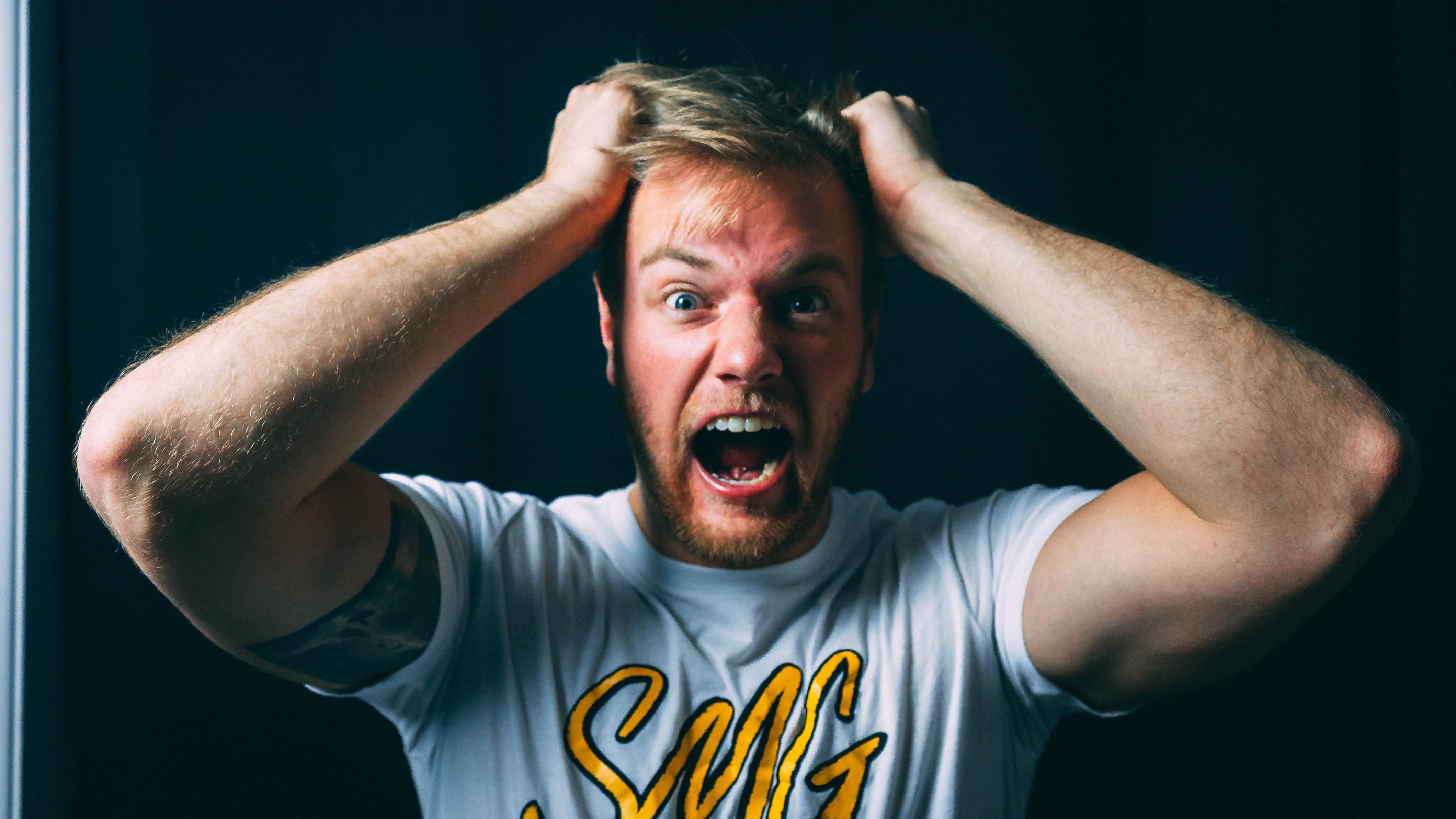 A bearded man with a distressed expression pulls his hair and opens his mouth, possibly yelling about spaghetti code, against a dark background. He wears a white T-shirt with yellow lettering.