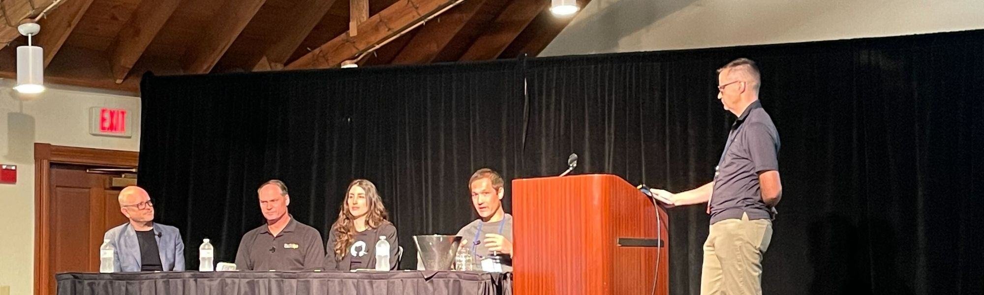 A panel of five people, four seated and one standing at a podium, engages in a discussion on generative AI. The room features wooden ceiling beams and a black curtain backdrop, with glasses and water bottles on the table, adding an aura of sophistication to the insightful exchange.