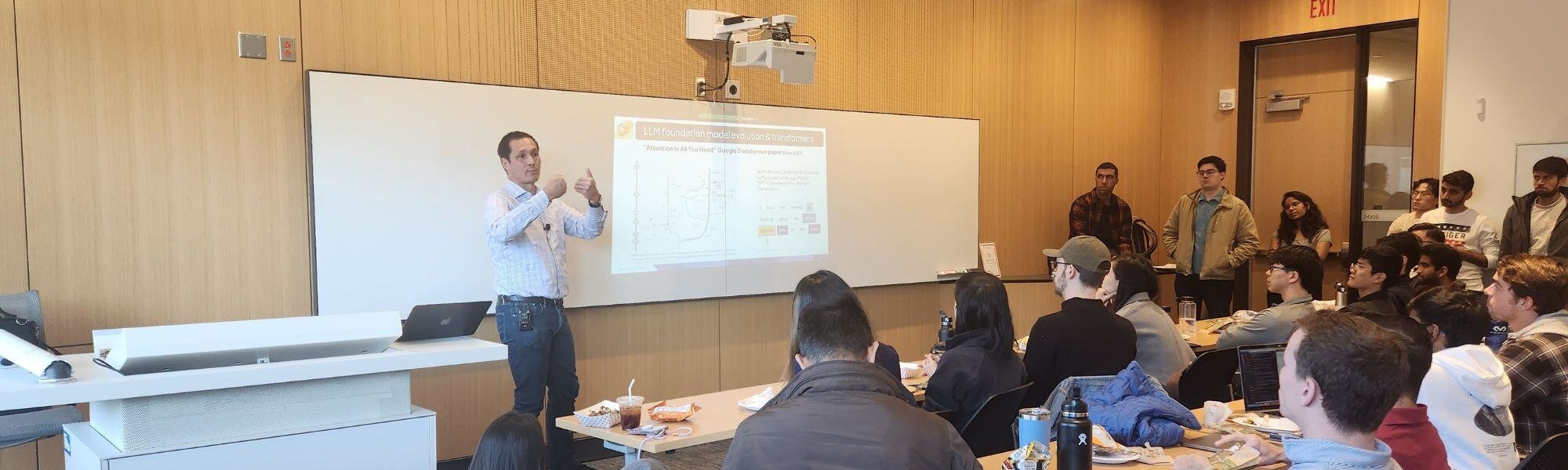 A man is giving a presentation on generative AI for business to a group in a classroom. Gesturing towards a projection screen displaying graphs and text, he captures the audience's attention, seated at tables and eagerly absorbing the innovative insights.
