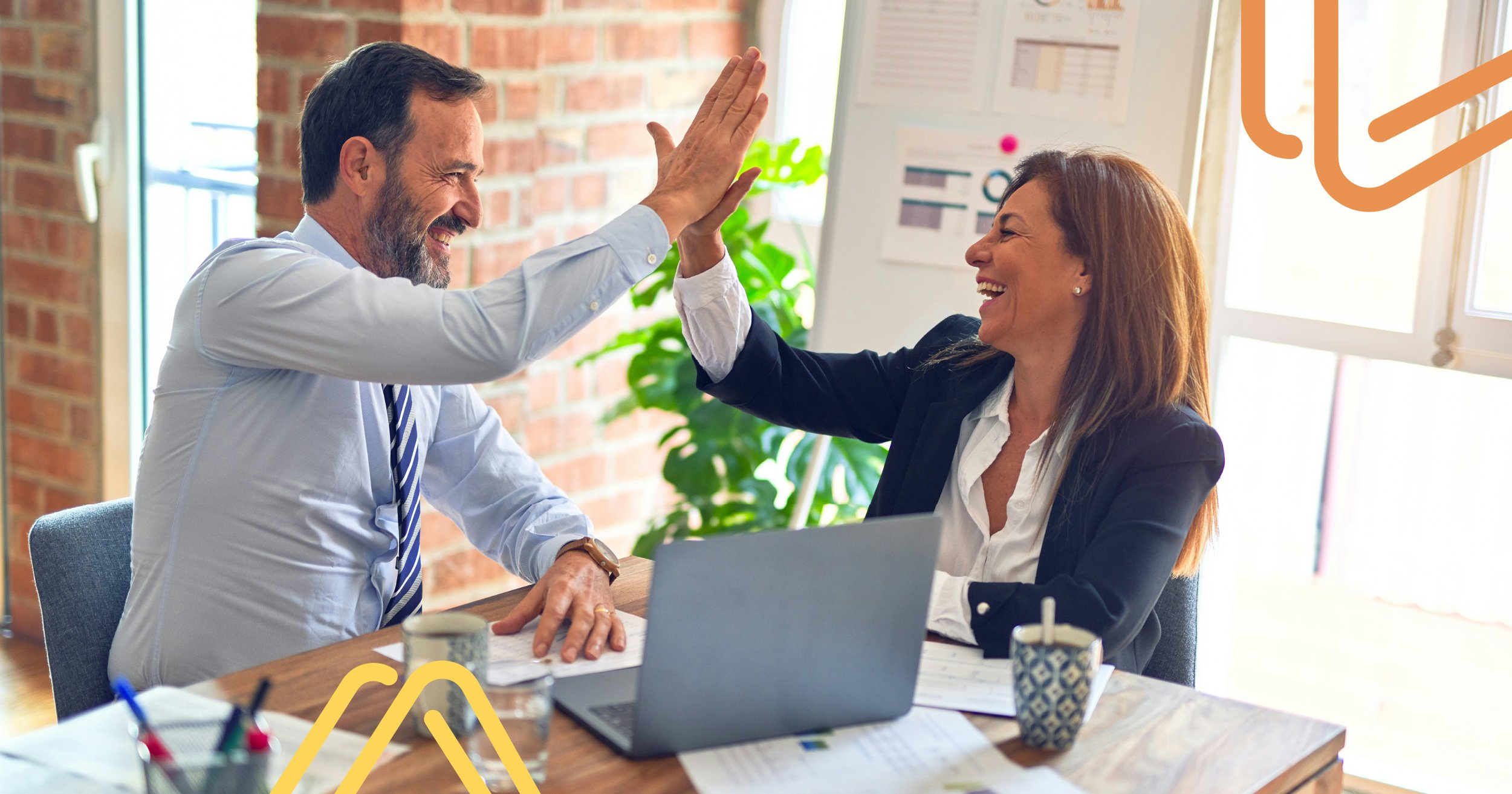 A man and woman in business attire share a high-five at a desk with a laptop and documents. They are smiling, suggesting a successful achievement. The background features a brick wall, window, and a whiteboard with charts.