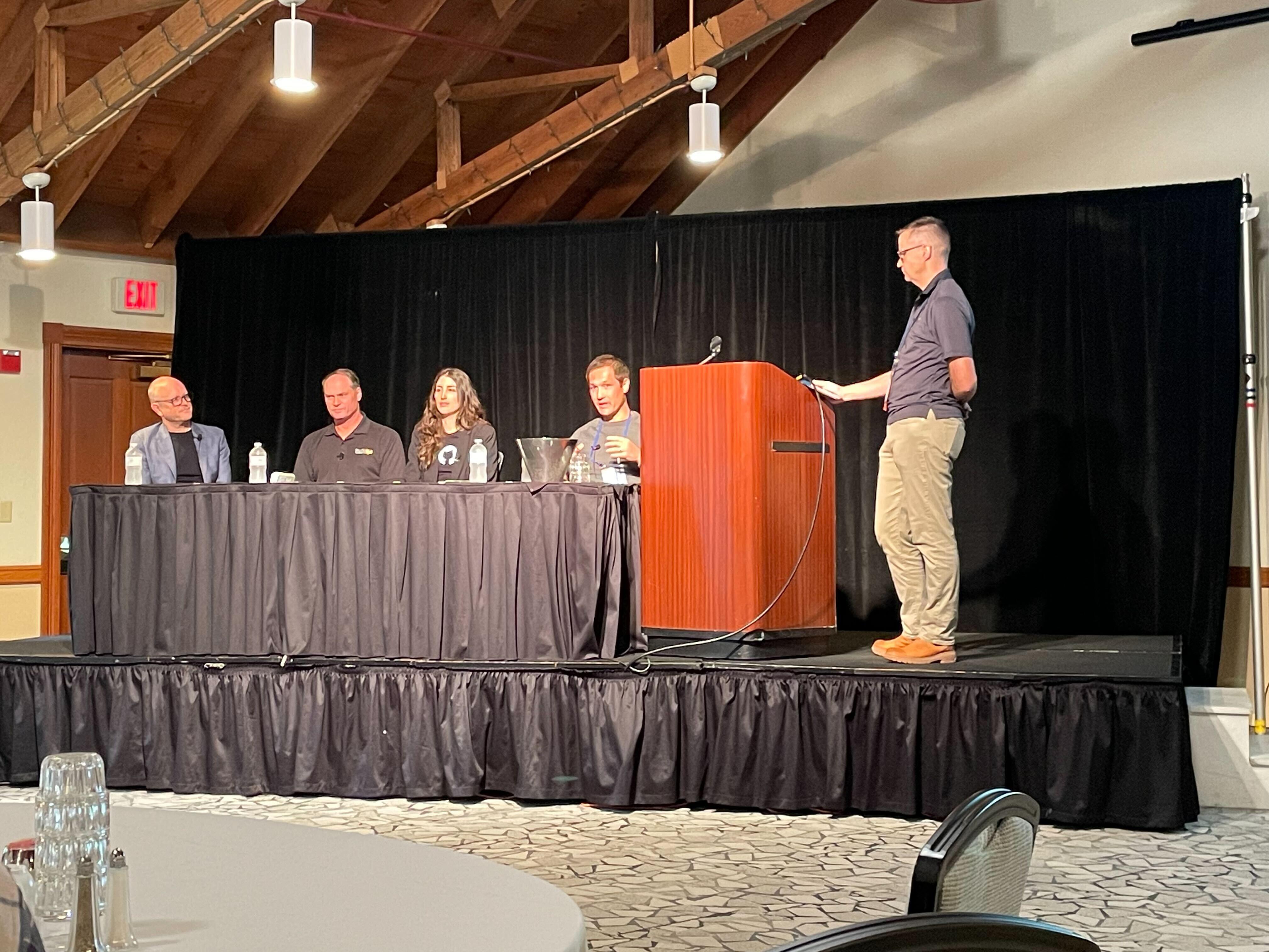 A panel of five people, four seated and one standing at a podium, engages in a discussion on generative AI. The room features wooden ceiling beams and a black curtain backdrop, with glasses and water bottles on the table, adding an aura of sophistication to the insightful exchange.